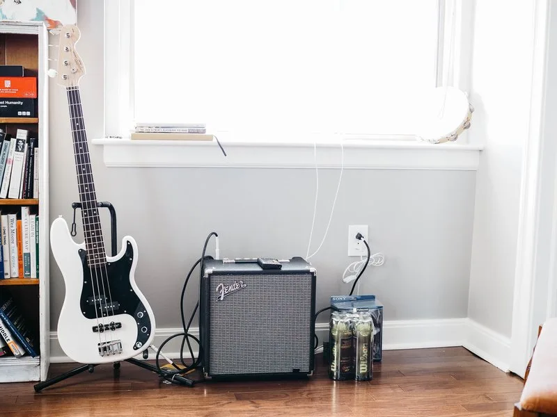A brightly lit room containing an electric guitar, Fender Guitar amp, and 2 unopenned packs of tennis balls